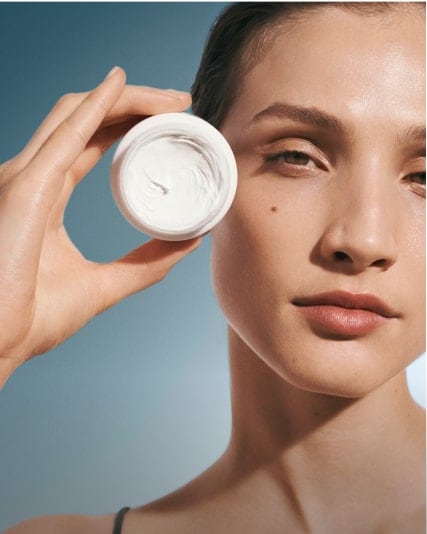 Model holding an open canister of cream up to the side of her face.